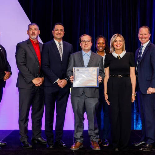 A group of seven people, dressed in business attire, stand on a stage between two flags. The person in the center is holding a framed certificate, and the group is posed in front of a dark curtain with geometric lighted shapes in the background.